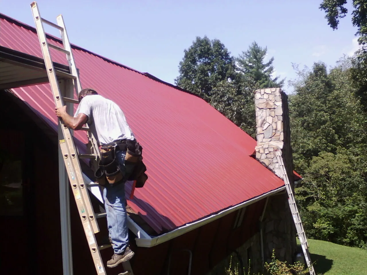 Roofer on a ladder beside a red metal roof and stone chimney — Soffit & Fascia Repair by Roofing Brothers in West Newton